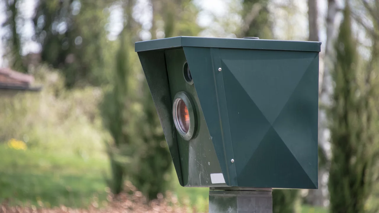 Eine Geschwindigkeitsmessanlage in Form eines grünen Kastens steht auf einem stabilen Pfosten. Sie ist auf einer Straße platziert und zeigt einen runden, orangefarbenen Belichtungspunkt. Der Hintergrund besteht aus Bäumen und Sträuchern.