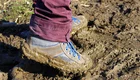 Ein Fuß steht in matschigem Boden. Der Schuh ist grau mit blauen Schnürsenkeln und ist mit Schlamm bedeckt. Der Hosenbund ist dunkelrot. Der Hintergrund zeigt eine grüne Wiese.
