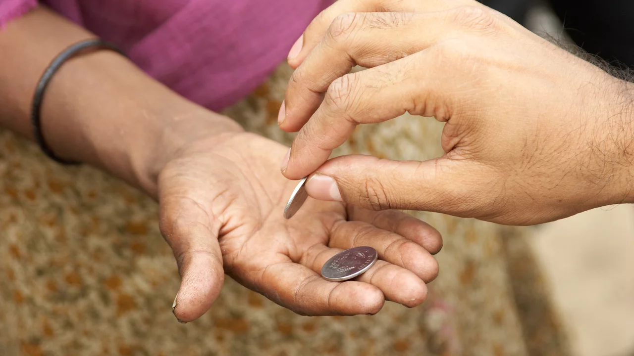 Eine Hand gibt eine Münze in die offene Hand einer anderen Person. Die Hände sind in Nahaufnahme zu sehen, mit Fokus auf den Moment der Übergabe der Münze.