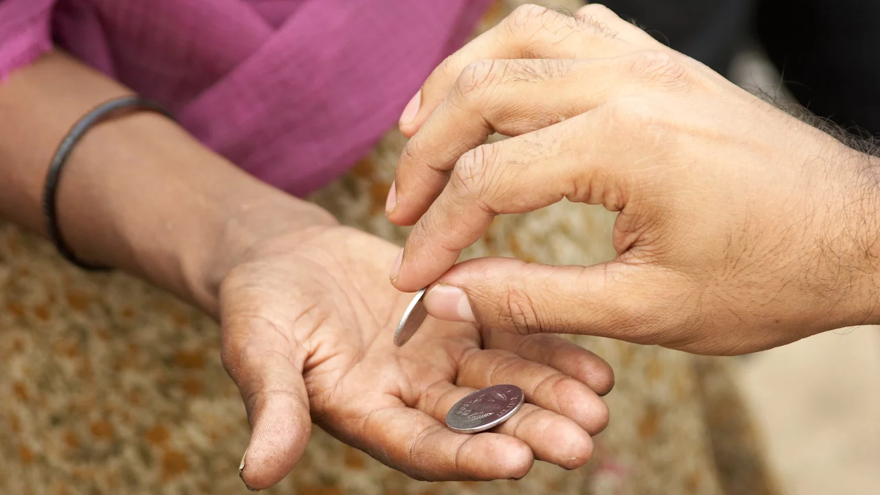Eine Hand gibt eine Münze in die offene Hand einer anderen Person. Die Hände sind in Nahaufnahme zu sehen, mit Fokus auf den Moment der Übergabe der Münze.