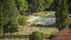 Eine belebte Parklandschaft an einem sonnigen Tag, in der viele Menschen auf Decken liegen, um sich zu entspannen. Ein Schwimmbecken ist im Hintergrund sichtbar, umgeben von Bäumen und Grünflächen. Die Atmosphäre wirkt lebhaft und einladend.