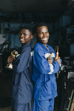 Zwei junge Männer in blauen Arbeitsanzügen stehen Rücken an Rücken in einer Werkstatt. Der linke Mann hält eine Bohrmaschine, der rechte eine Schraubendreher. Beide lächeln und strahlen Selbstbewusstsein aus, während Werkzeuge und Regale im Hintergrund zu sehen sind.