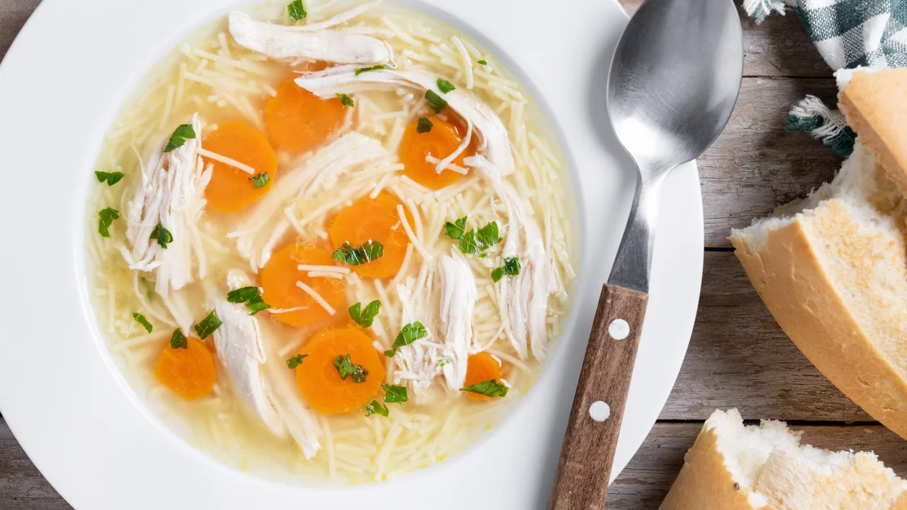 Eine Schüssel Hühnersuppe mit feinen Nudeln, Stückchen von Hähnchenfleisch und Karotten. Die Suppe ist mit frischen Kräutern garniert. Neben der Schüssel liegt ein Löffel und einige Brotscheiben auf einem Holztisch.