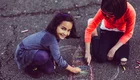 Zwei Kinder sitzen auf dem Asphalt und zeichnen mit Kreide. Ein Mädchen lächelt fröhlich, während sie an einer bunten Zeichnung arbeitet. Das andere Kind konzentriert sich ebenfalls auf das Malen. Die Szene vermittelt Freude und Kreativität beim Spielen im Freien.