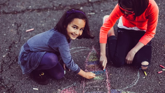 Zwei Kinder sitzen auf dem Asphalt und zeichnen mit Kreide. Ein Mädchen lächelt fröhlich, während sie an einer bunten Zeichnung arbeitet. Das andere Kind konzentriert sich ebenfalls auf das Malen. Die Szene vermittelt Freude und Kreativität beim Spielen im Freien.