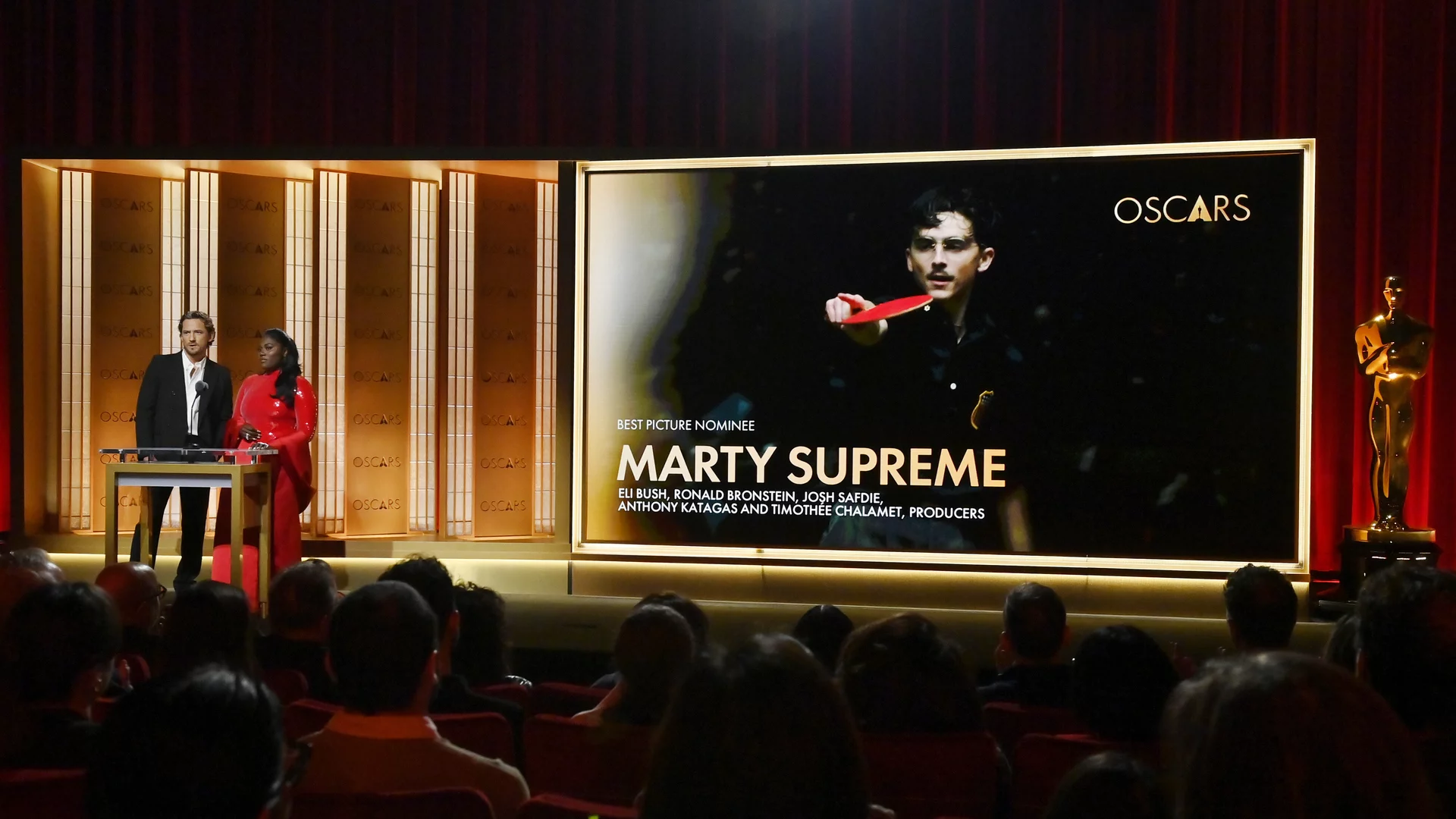 Auf der Bühne bei den Oscars stehen zwei Moderatoren. Im Hintergrund wird die Nominierung für "Best Picture" angezeigt: "Marty Supreme". Es sind die Namen der Produzenten aufgelistet. Das Publikum im Vordergrund lauscht aufmerksam.
