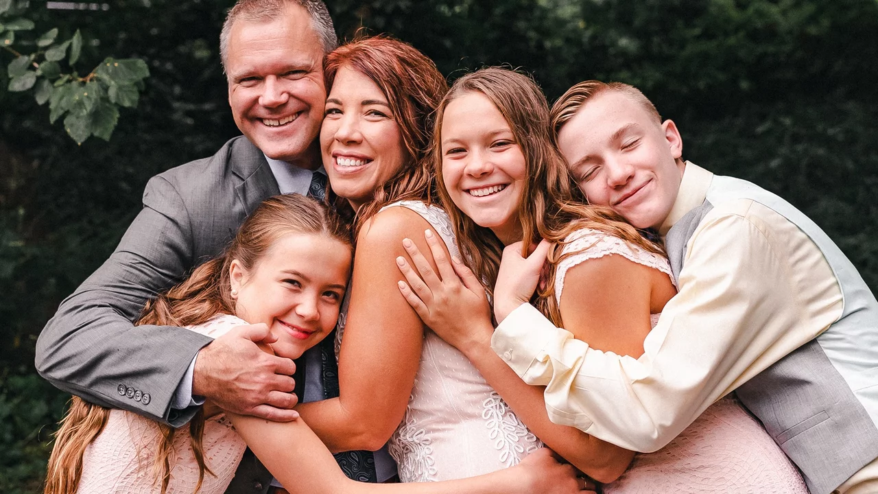 Ein lächelndes Familienporträt zeigt einen Vater und eine Mutter, umgeben von drei glücklichen Kindern. Alle umarmen sich, tragen festliche Kleidung und wirken liebevoll verbunden. Die Szene vermittelt Wärme und Zusammenhalt in einem natürlichen Hintergrund.