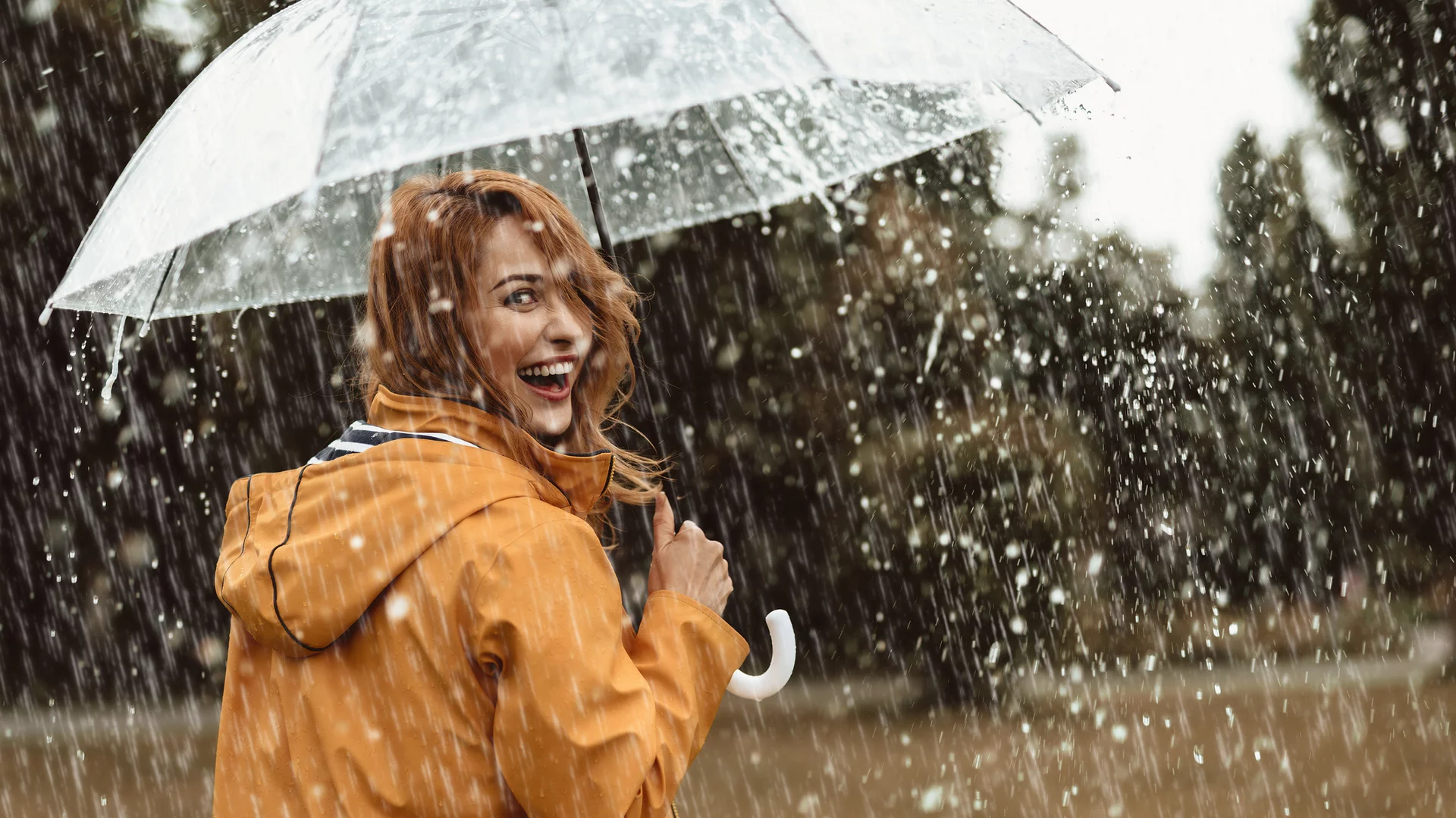 Eine lächelnde Frau mit roten Haaren trägt einen gelben Regenmantel und hält einen durchsichtigen Regenschirm. Sie steht im Regen, während Wasser tropft und die Umgebung unscharf wirkt. Ihre fröhliche Ausstrahlung vermittelt Freude trotz des regnerischen Wetters.