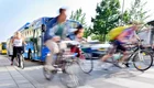 Auf der Straße überqueren mehrere Radfahrer in Bewegung einen Zebrastreifen, während ein blauer Bus im Hintergrund sichtbar ist. Die Szene vermittelt ein Gefühl von lebhaftem Verkehr und urbaner Aktivität.