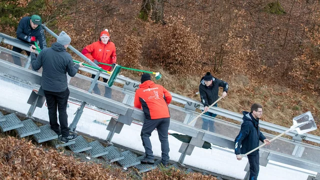 Sechs Personen arbeiten zusammen, um die Schanze in Willingen für den Weltcup im Skispringen zu reinigen oder Instand zu halten. Sie verwenden verschiedene Werkzeuge und stehen auf einer schneebedeckten Rampe. Im Hintergrund sind Bäume und abgestorbene Blätter sichtbar.