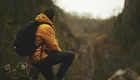 Ein Mann sitzt auf einem Felsen und schaut auf eine weite, bewaldete Schlucht. Er trägt eine orangefarbene Jacke und einen schwarzen Rucksack. Die Umgebung wirkt ruhig und natürlich, mit sanften Hügeln im Hintergrund.