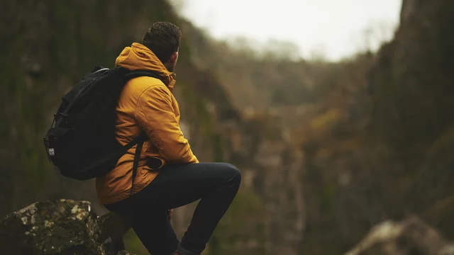 Ein Mann sitzt auf einem Felsen und schaut auf eine weite, bewaldete Schlucht. Er trägt eine orangefarbene Jacke und einen schwarzen Rucksack. Die Umgebung wirkt ruhig und natürlich, mit sanften Hügeln im Hintergrund.