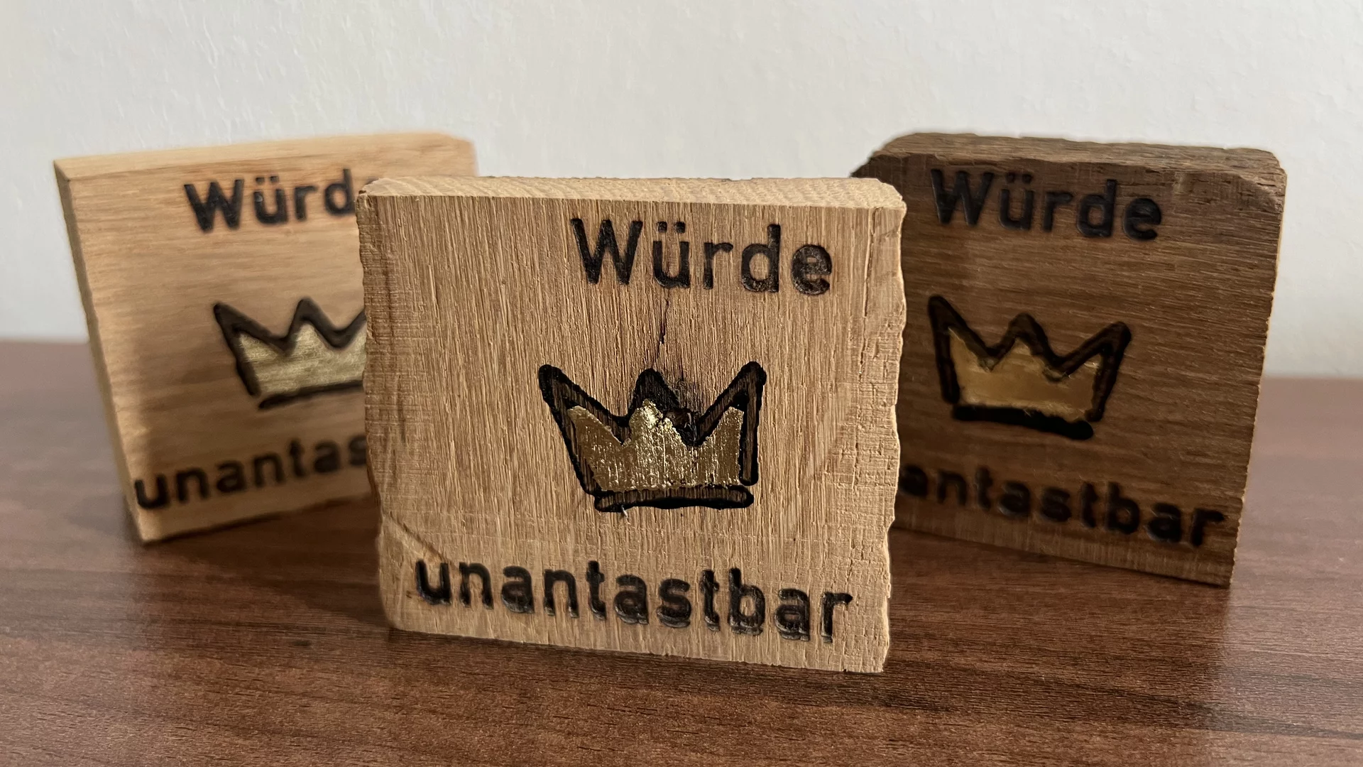 Drei Holzbretter liegen auf einer Oberfläche. Auf jedem Brett steht die Inschrift „Würde unantastbar“ in schwarzer Schrift, begleitet von einer stilisierten Krone in Gold. Die Bretter haben unterschiedliche Holzfarben, die von hell bis dunkel reichen.