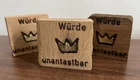 Drei Holzbretter liegen auf einer Oberfläche. Auf jedem Brett steht die Inschrift „Würde unantastbar“ in schwarzer Schrift, begleitet von einer stilisierten Krone in Gold. Die Bretter haben unterschiedliche Holzfarben, die von hell bis dunkel reichen.