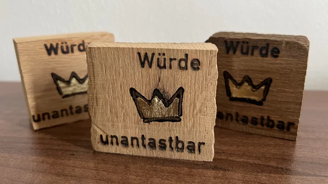 Drei Holzbretter liegen auf einer Oberfläche. Auf jedem Brett steht die Inschrift „Würde unantastbar“ in schwarzer Schrift, begleitet von einer stilisierten Krone in Gold. Die Bretter haben unterschiedliche Holzfarben, die von hell bis dunkel reichen.