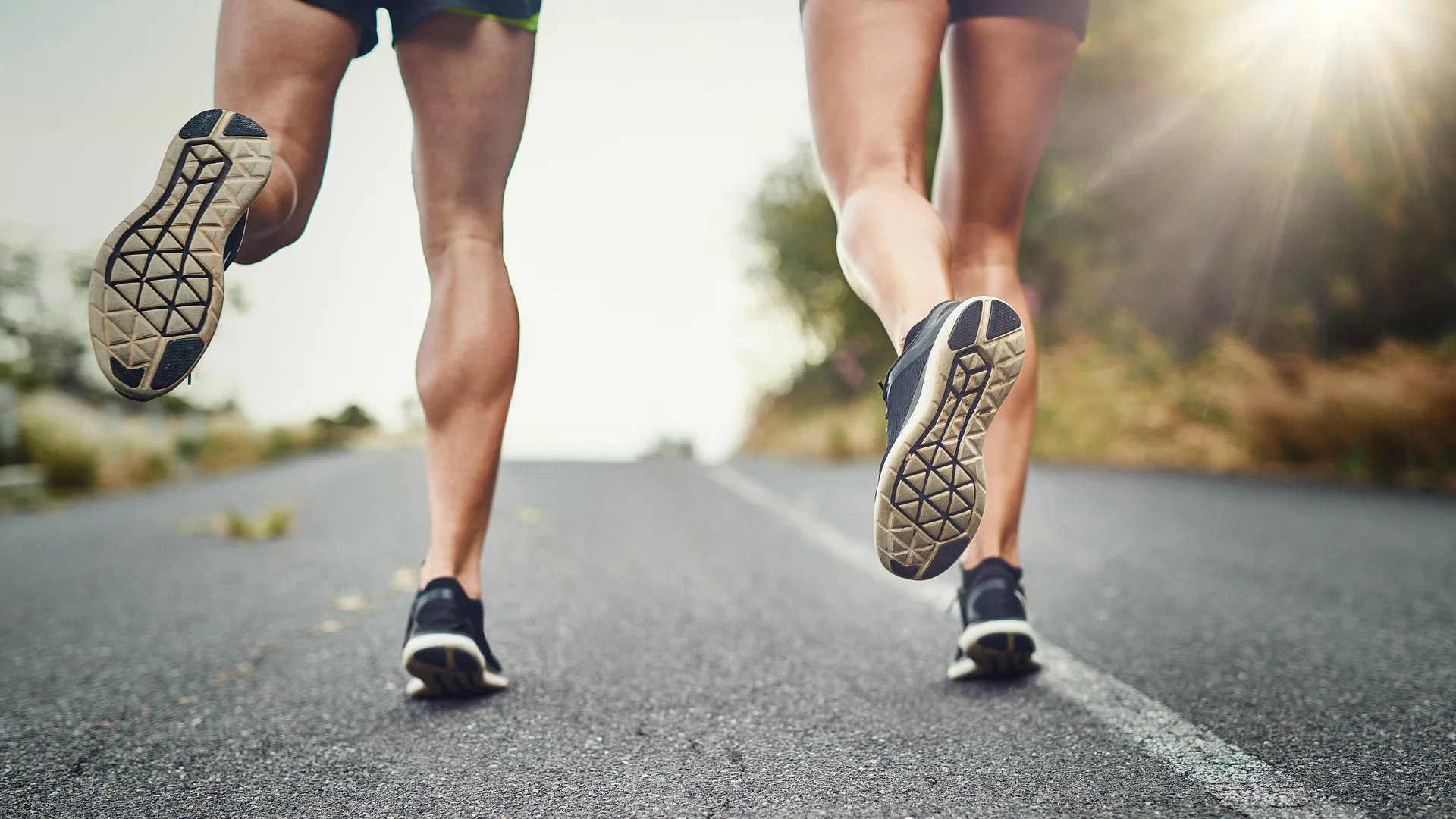 Zwei Personen joggen auf einer Straße, die Kamera zeigt hauptsächlich ihre Beine und Schuhe. Die Umgebung ist grün und etwas verschwommen, mit einem sanften Lichtschein, der den Eindruck eines sonnigen Tages vermittelt.