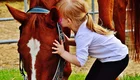 Ein kleines Mädchen kniet neben einem braunen Pferd und küsst sanft seine Schnauze. Das Mädchen trägt ein weißes Oberteil und schwarze Leggings, während das Pferd ruhig steht. Die Szene vermittelt eine liebevolle Verbindung zwischen dem Kind und dem Tier.