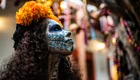 Eine Frau mit aufwendig geschminktem Gesicht, das einem Totenkopf ähnelt, trägt einen Haarreif mit orangefarbenen Blumen. Sie blickt zur Seite, während farbenfrohe Dekorationen im Hintergrund erkennbar sind. Die Szene vermittelt eine festliche Atmosphäre.