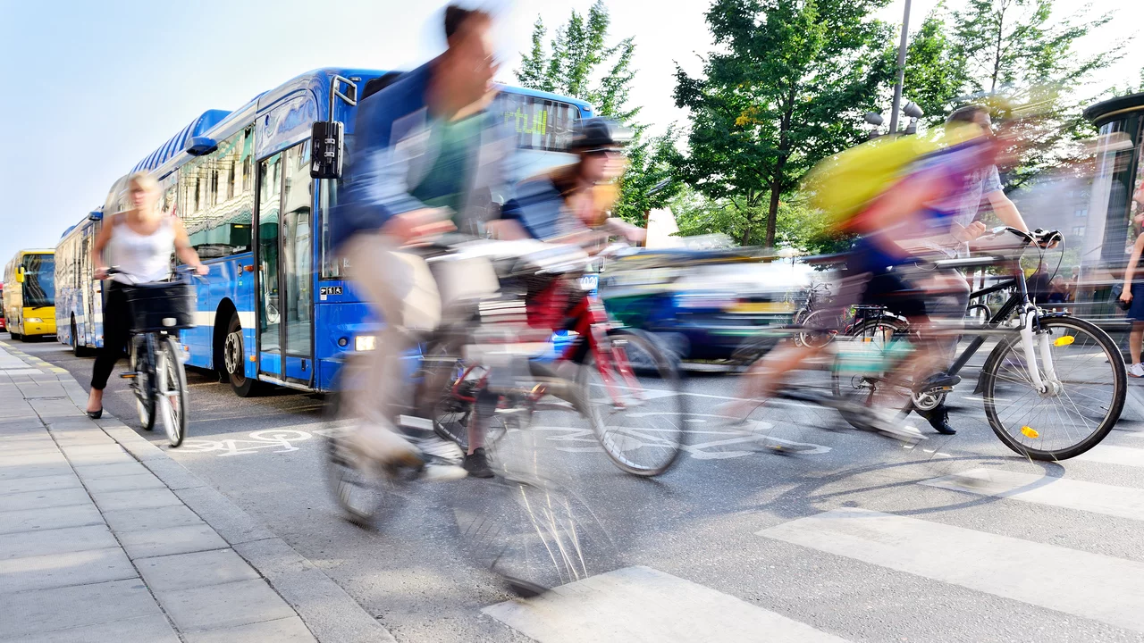 Zwei Radfahrer fahren auf einem Zebrastreifen, während ein Bus vorbeifährt. Die Szene ist dynamisch, mit Bewegungsunschärfe, die die Geschwindigkeit der Radfahrer verstärkt. Im Hintergrund sind Bäume und weitere Fahrzeuge zu sehen.