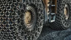 Nahaufnahme von zwei schwer beladenen Reifen eines Fahrzeugs. Die Reifen tragen Schneeketten, die eine robuste Struktur aufweisen. Der Boden ist mit grobem Material bedeckt, was auf eine raue Umgebung hinweist.