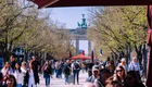 Ein belebter Boulevard führt zum Brandenburger Tor, umgeben von Bäumen und flanierenden Menschen. In der Ferne ist eine zentrale rote Pagode zu sehen, die als Treffpunkt dient. Die Szene strahlt lebendige Aktivität und Geselligkeit aus.