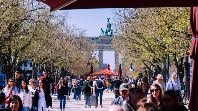 Ein belebter Boulevard führt zum Brandenburger Tor, umgeben von Bäumen und flanierenden Menschen. In der Ferne ist eine zentrale rote Pagode zu sehen, die als Treffpunkt dient. Die Szene strahlt lebendige Aktivität und Geselligkeit aus.
