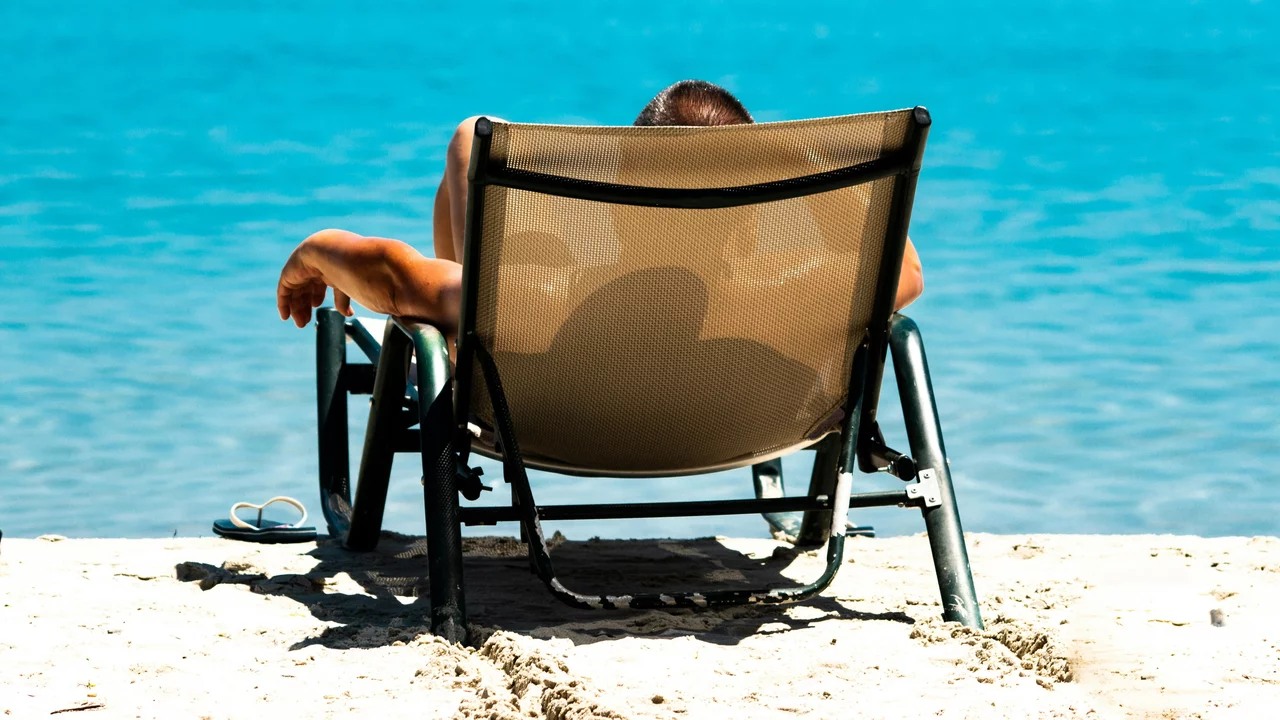 Eine Person sitzt entspannt auf einem Liegestuhl am Strand, mit dem Rücken zur Kamera. Im Hintergrund ist das türkisfarbene Wasser des Meeres sichtbar, das sanft an den Sandstrand plätschert. Die Szene vermittelt ein Gefühl von Ruhe und Entspannung.
