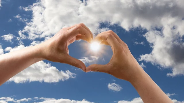 Zwei Hände formen ein Herzsymbol vor einem strahlend blauen Himmel mit Wolken. In der Mitte des Herzens scheint die Sonne, was eine warme und positive Stimmung vermittelt.