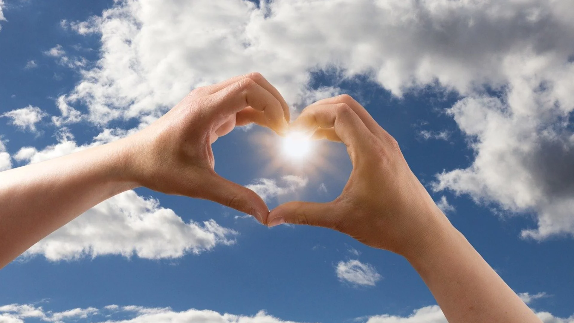 Zwei Hände formen ein Herzsymbol vor einem strahlend blauen Himmel mit Wolken. In der Mitte des Herzens scheint die Sonne, was eine warme und positive Stimmung vermittelt.