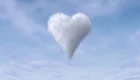 Eine herzförmige Wolke schwebt vor einem blauen Himmel. Die Wolke ist weich und flauschig, umgeben von leichter Bewölkung, die die romantische und verspielte Stimmung unterstreicht.