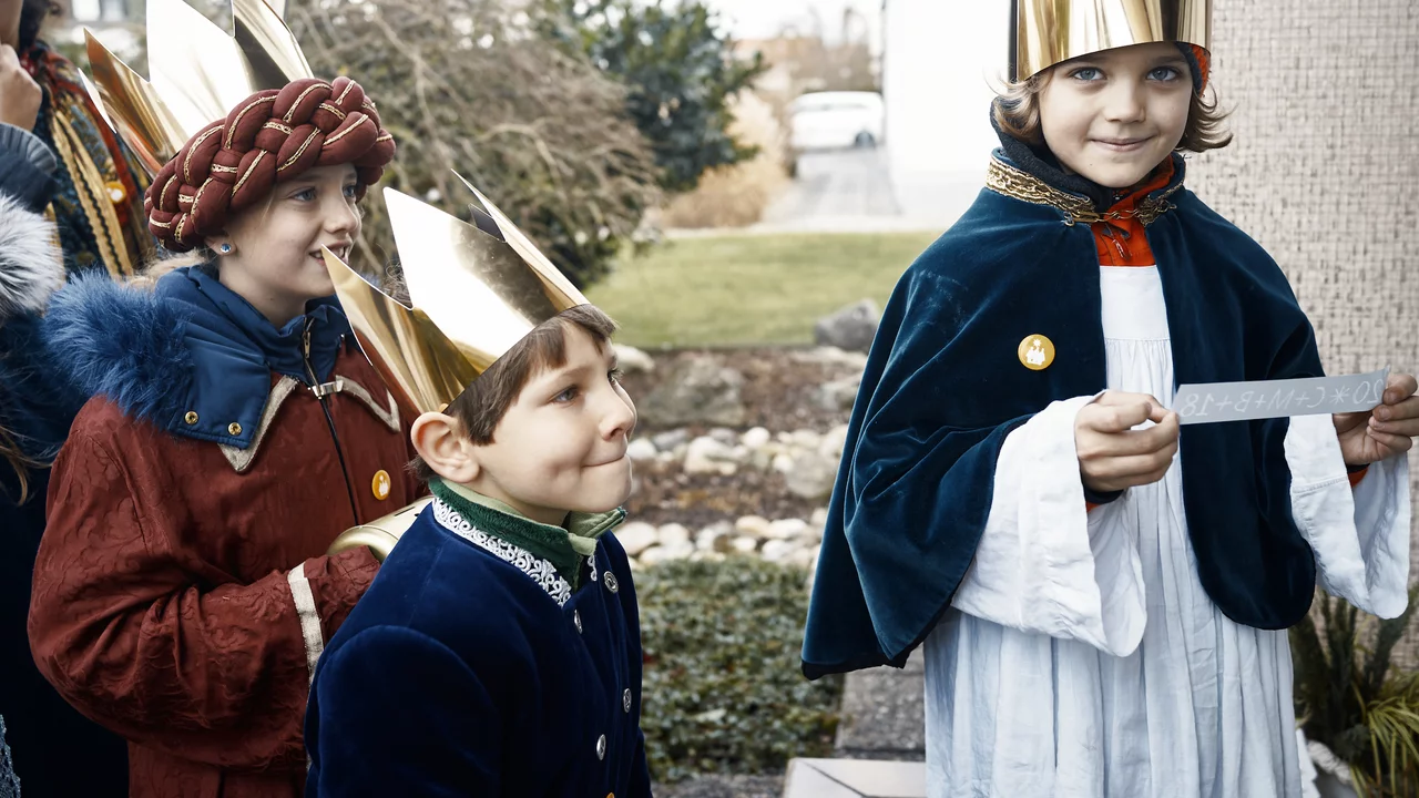 Drei Kinder in bunten Kostümen und goldenen Kronen stehen vor einer Haustür. Eines der Kinder hält ein Stück Papier in der Hand. Sie lächeln und wirken fröhlich, während sie auf den Eingang schauen.
