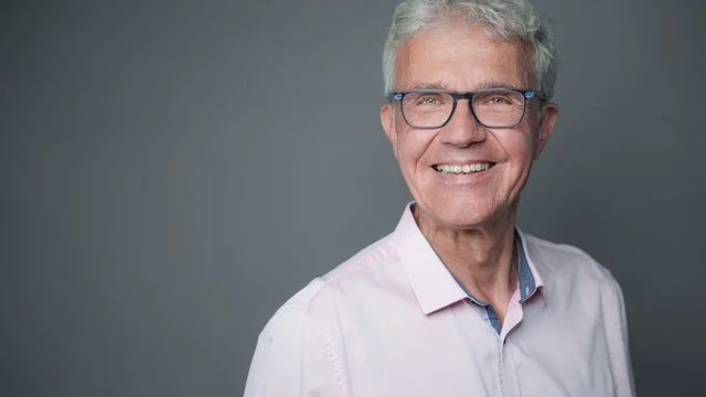 Ein älterer Mann mit grauen Haaren und Brille lächelt freundlich. Er trägt ein helles Hemd und steht vor einem neutralen Hintergrund. Sein Ausdruck vermittelt Wärme und Offenheit.