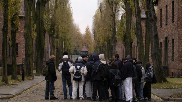 Eine Gruppe von Jugendlichen steht in einer Allee, umgeben von Bäumen und den Gebäuden von Auschwitz I. Die Szene vermittelt eine nachdenkliche Atmosphäre an diesem Ort des Gedenkens und der Bildung über die Geschichte. Das Wetter ist trüb und die Landschaft wirkt melancholisch.