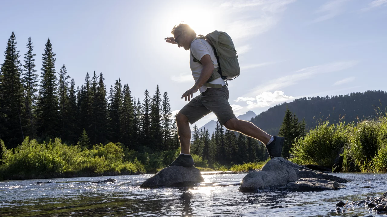 Ein Wanderer mit Rucksack springt von Stein zu Stein über einen Fluss. Im Hintergrund sind grüne Bäume und Berge zu sehen, während die Sonne über der Szenerie strahlt und ein schönes Licht erzeugt.