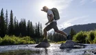 Ein Wanderer mit Rucksack springt von Stein zu Stein über einen Fluss. Im Hintergrund sind grüne Bäume und Berge zu sehen, während die Sonne über der Szenerie strahlt und ein schönes Licht erzeugt.
