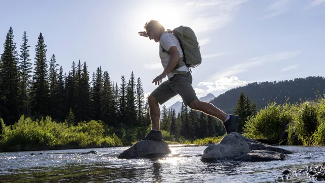 Ein Wanderer mit Rucksack springt von Stein zu Stein über einen Fluss. Im Hintergrund sind grüne Bäume und Berge zu sehen, während die Sonne über der Szenerie strahlt und ein schönes Licht erzeugt.