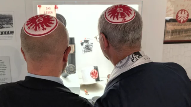 Zwei Männer mit Kippot auf dem Kopf stehen vor einem Vitrinenausstellungsstück. Sie betrachten darin Erinnerungsstücke und Informationen, die die Themen Leben und Geschichte behandeln. Die Kippot zeigen die Farben und das Wappen eines Fußballvereins.