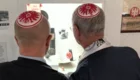 Zwei Männer mit Kippot auf dem Kopf stehen vor einem Vitrinenausstellungsstück. Sie betrachten darin Erinnerungsstücke und Informationen, die die Themen Leben und Geschichte behandeln. Die Kippot zeigen die Farben und das Wappen eines Fußballvereins.