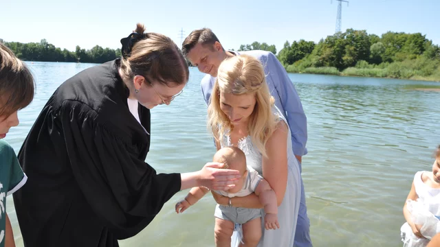 Eine Tauffeier am Wasser. Eine Frau in einem Talar tauft ein Baby, während die Eltern, ein Mann und eine Frau, aufmerksam zuschauen. Im Hintergrund sind Bäume und eine ruhige Wasserfläche zu sehen. Es ist ein sonniger Tag.