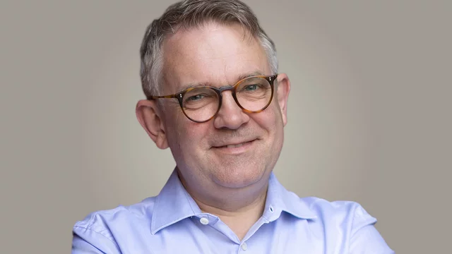 Ein lächelnder Mann mit Brille und grau meliertem Haar trägt ein hellblaues Hemd. Er hat die Arme verschränkt und sieht freundlich aus. Der Hintergrund ist in neutralen Farbtönen gehalten.