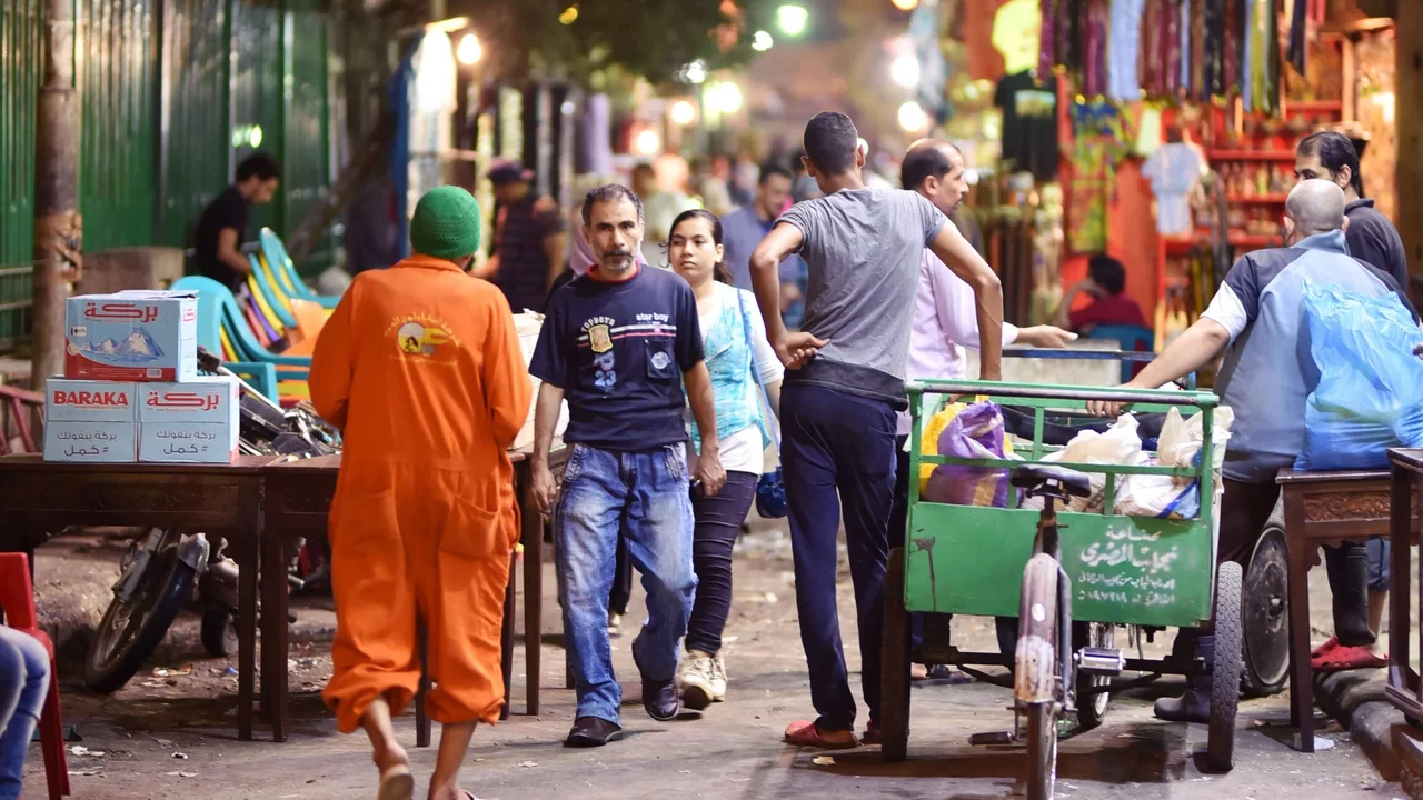 Eine belebte Straße in einer Stadt bei Nacht. Menschen gehen umher, einige unterhalten sich. Verkaufsstände sind im Hintergrund sichtbar. Eine Person trägt einen orangenen Overall. Ein Handwagen steht auf dem Bürgersteig. Die Szene vermittelt eine lebendige, geschäftige Atmosphäre.