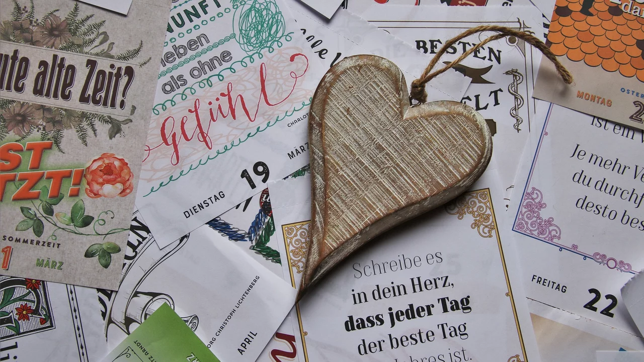Ein hölzernes Herz liegt auf einem bunten Hintergrund aus verschiedenen Papieren und Karten mit inspirierenden Sprüchen und Datumshinweisen. Das Herz symbolisiert Liebe und Wertschätzung, während die Papiere positive Botschaften vermitteln.