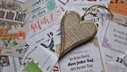 Ein hölzernes Herz liegt auf einem bunten Hintergrund aus verschiedenen Papieren und Karten mit inspirierenden Sprüchen und Datumshinweisen. Das Herz symbolisiert Liebe und Wertschätzung, während die Papiere positive Botschaften vermitteln.