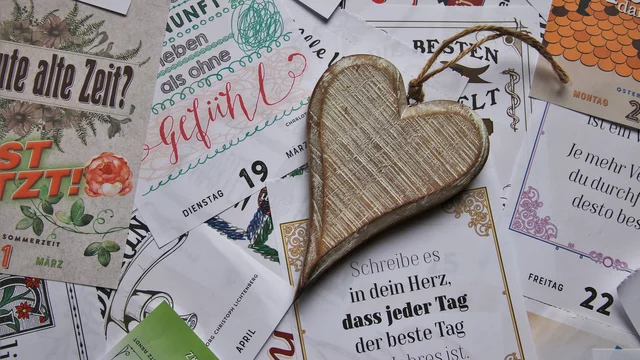 Ein hölzernes Herz liegt auf einem bunten Hintergrund aus verschiedenen Papieren und Karten mit inspirierenden Sprüchen und Datumshinweisen. Das Herz symbolisiert Liebe und Wertschätzung, während die Papiere positive Botschaften vermitteln.