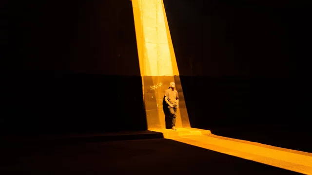 Eine Person steht in einem dunklen Raum, beleuchtet von einem hellen, schmalen Lichtstrahl. Die Atmosphäre wirkt dramatisch und kontrastreich, mit starken Schatten und warmen Lichttönen.