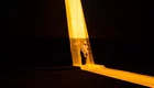 Eine Person steht in einem dunklen Raum, beleuchtet von einem hellen, schmalen Lichtstrahl. Die Atmosphäre wirkt dramatisch und kontrastreich, mit starken Schatten und warmen Lichttönen.