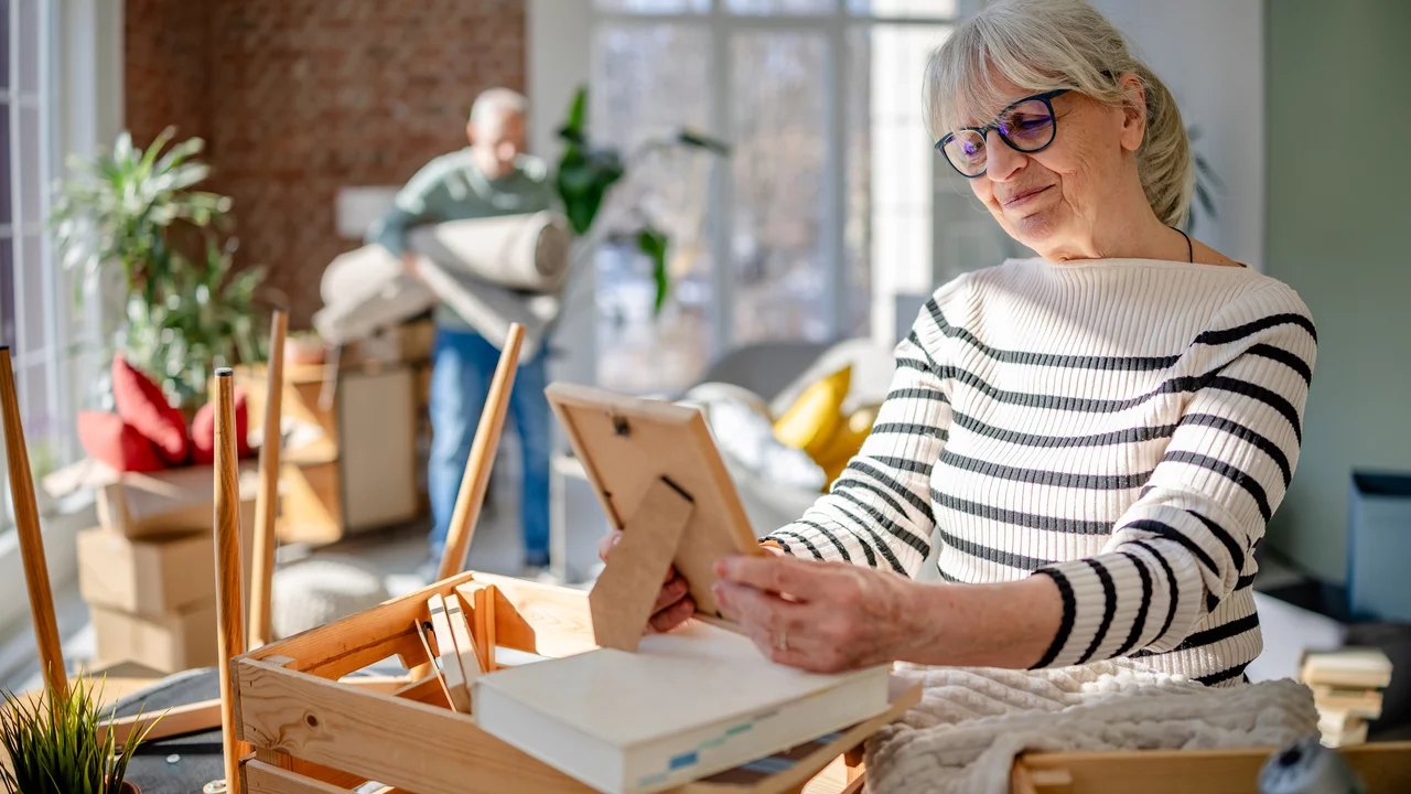 Eine ältere Frau mit grauen Haaren und Brille sitzt an einem Tisch und betrachtet ein Foto in einem Bilderrahmen. Im Hintergrund ist ein älterer Mann zu sehen, der einen Teppich in den Raum bringt. Der Raum wirkt hell und freundlich, mit Umzugskisten und Pflanzen.