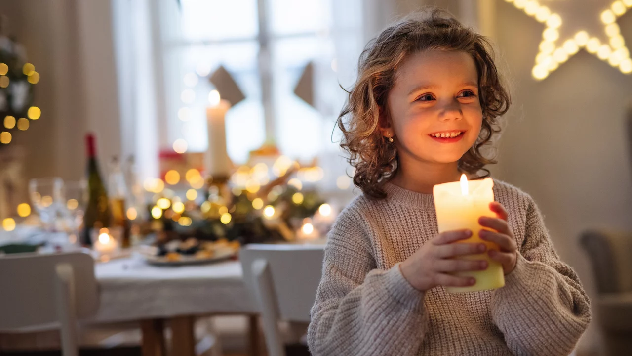 Ein Kind mit lockigem Haar lächelt und hält eine leuchtende Kerze in der Hand. Im Hintergrund ist ein festlich dekorierter Tisch mit Lichtern und Weihnachtsdekoration sichtbar. Die Atmosphäre ist warm und einladend.