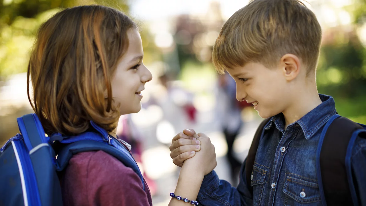 Zwei Kinder, ein Junge und ein Mädchen, stehen beieinander und lächeln einander an. Sie halten sich an den Händen, wobei sie sich fest zudrücken. Im Hintergrund sind verschwommene Silhouetten von anderen Kindern und Bäumen zu sehen. Die Szene strahlt Freundschaft und Freude aus.