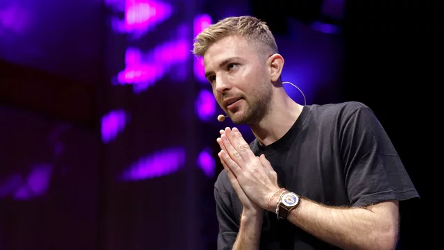 Ein Mann - Fußball Weltmeister Christoph Kramer -  in einem schwarzen T-Shirt steht auf einer Bühne und spricht. Er hat die Hände in einer betenden Geste zusammengelegt und schaut aufmerksam. Im Hintergrund sind diffuse, lila Lichtmuster zu sehen, die eine lebendige Atmosphäre schaffen.